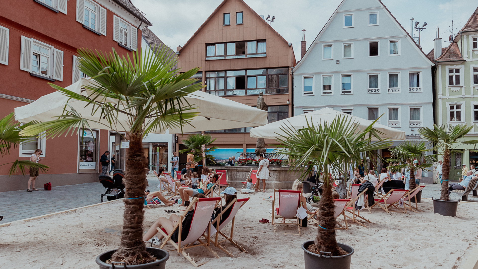 Städtischer Sandstrand mit Liegestühlen, Sonnenschirmen und Palmen in großen Kübeln, umgeben von bunten Fachwerkhäusern; Menschen entspannen in sommerlicher Atmosphäre mitten in der Innenstadt.