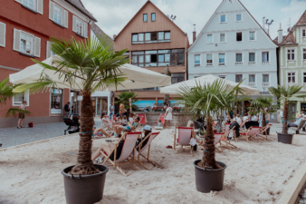Städtischer Sandstrand mit Liegestühlen, Sonnenschirmen und Palmen in großen Kübeln, umgeben von bunten Fachwerkhäusern; Menschen entspannen in sommerlicher Atmosphäre mitten in der Innenstadt.